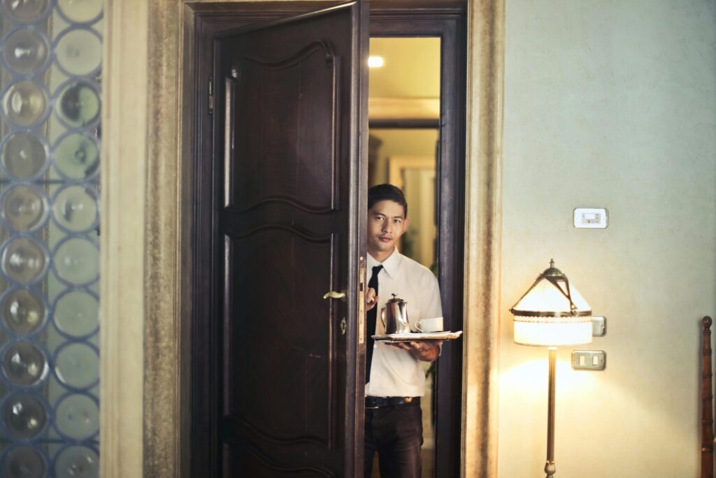 Young ethnic male room service waiter carrying tray with coffee pot while entering hotel room with stylish vintage interior