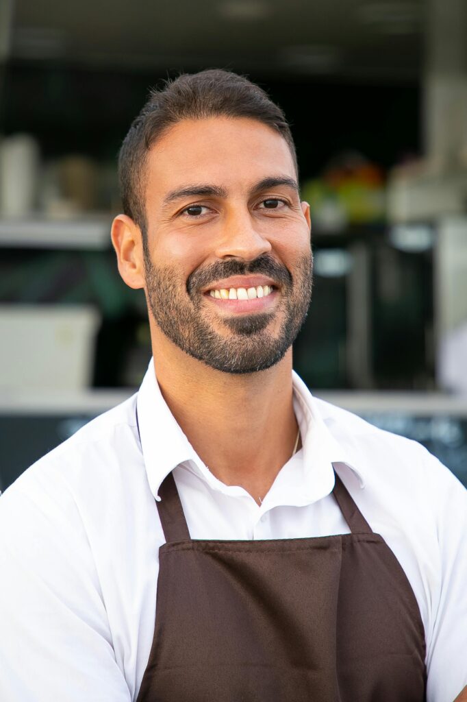 Smiling male café worker in apron, standing confidently outdoors in a business setting.